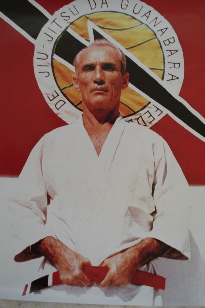 Portrait d'Hélio Gracie âgé, en kimono blanc, assis devant un emblème de ju-jitsu