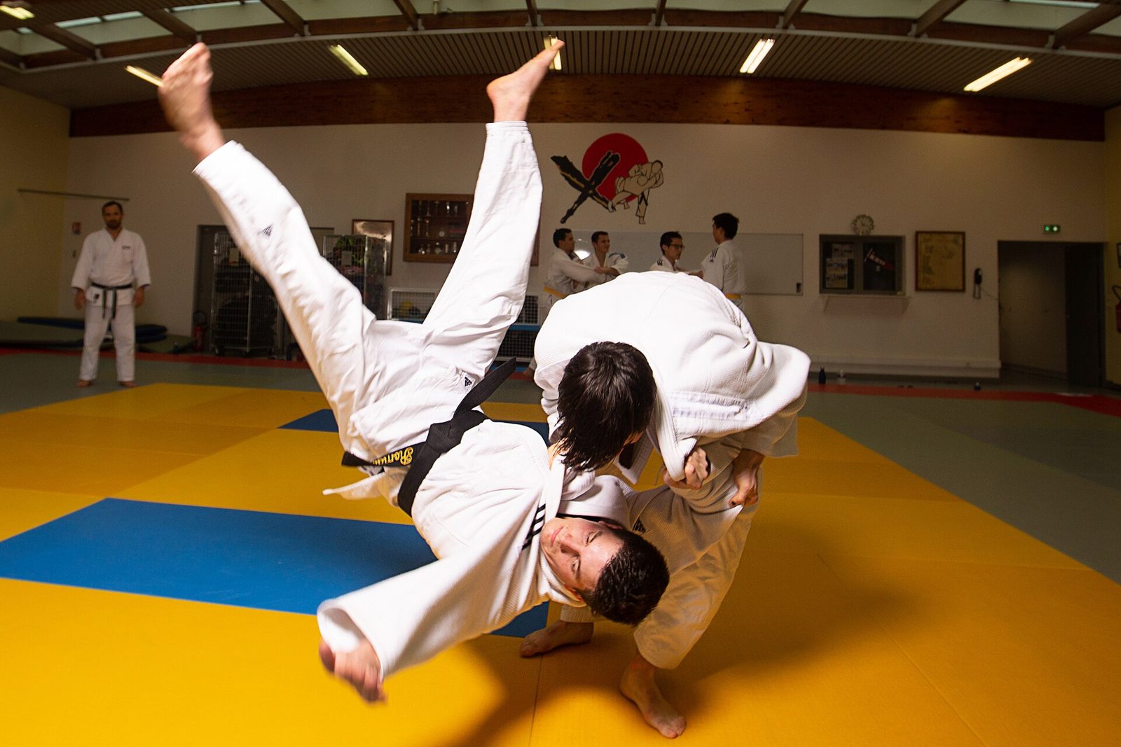 Un judoka en kimono blanc projeté en l'air par son partenaire sur un tatami jaune et bleu