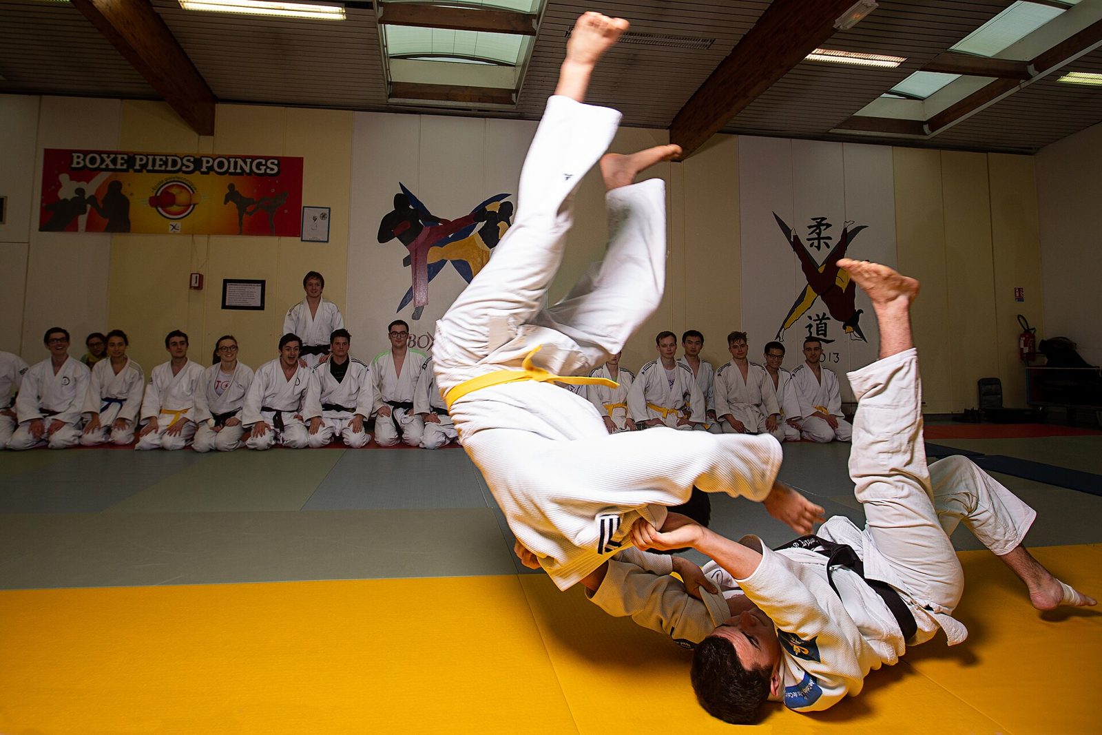 Projection spectaculaire pendant un entraînement, sous le regard de judokas assis en bord de tatami